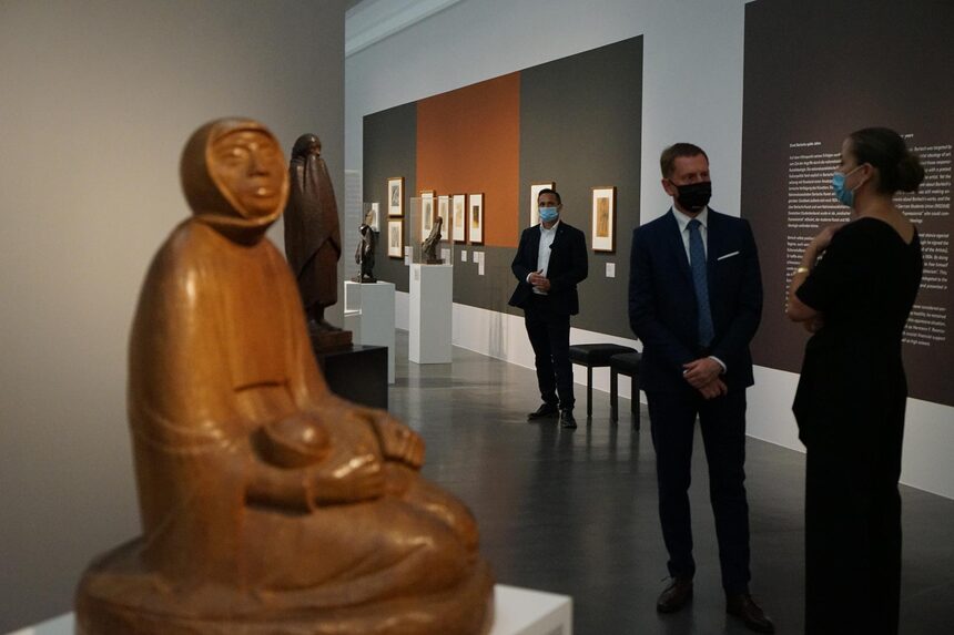 Ein Mann unterhält sich in einem Museumssaal mit einer Frau. Im Vordergrund steht eine Holzskulptur.