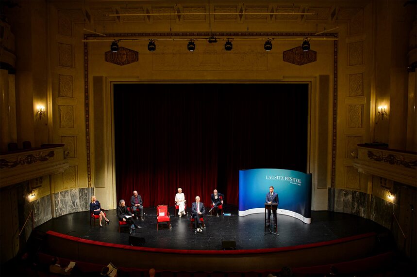 Sechs Menschen sitzen auf einer Theaterbühne vor einem roten Vorhang. Ein Mann neben ihnen hinter einem Rednerpult.