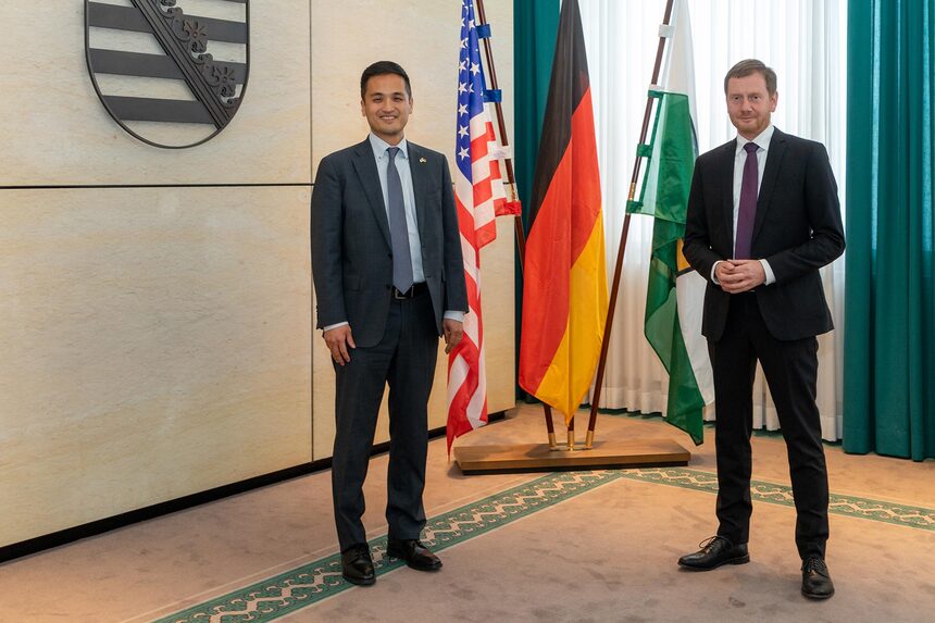 Zwei Männer stehen vor der deutschen, der sächsischen und der US-amerikanischen Fahne.