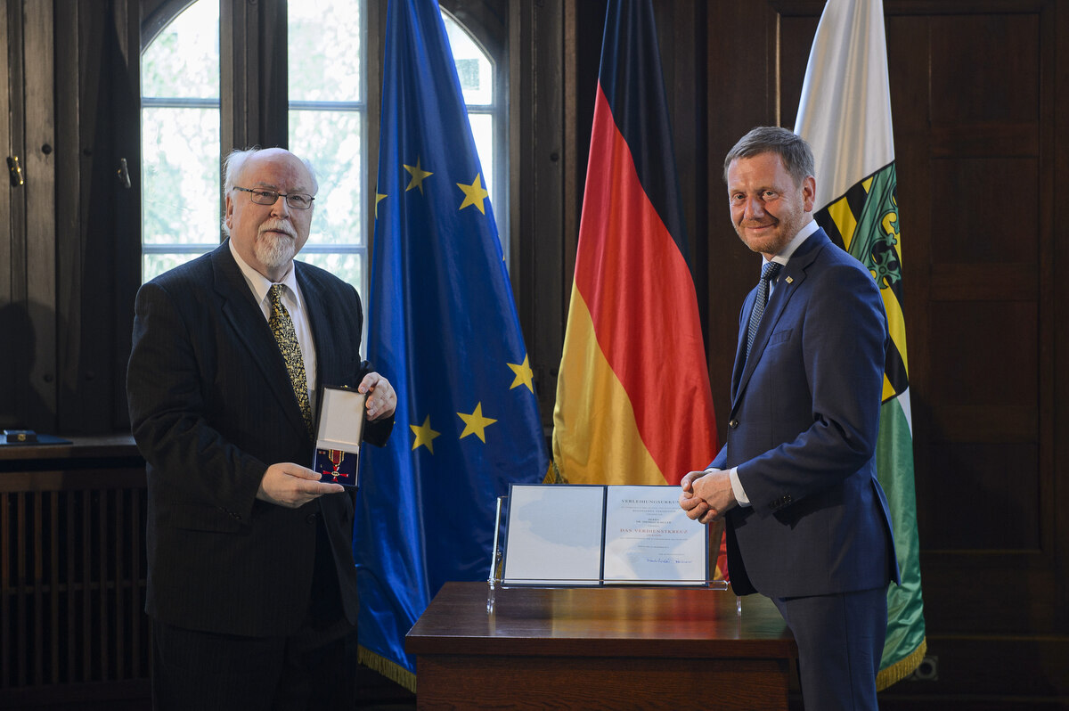 Zwei Männer stehen neben einem Pult. Einer hält einen Orden in der Hand.