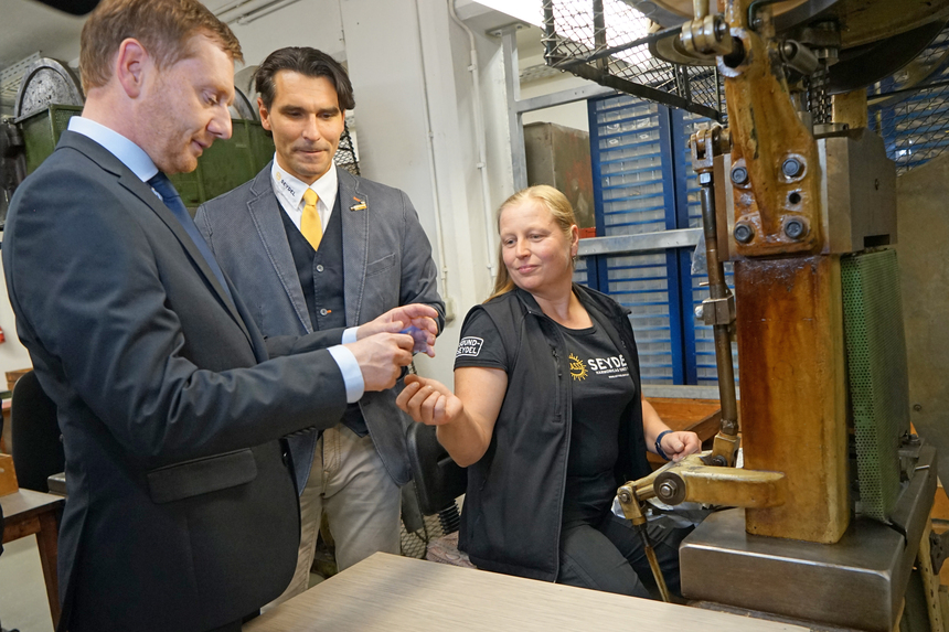 Eine Frau reicht einem Mann ein gepresstes Metallstück. Daneben steht ein weterer Mann und schaut zu.