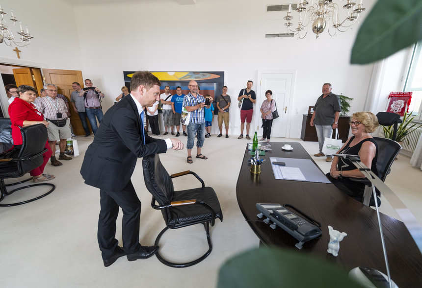 Ein Büro das von vielen Besuchern besichtigt wird. An einem Schreibtisch sitzt eine Frau, davor steht eine Mann und schaut sie an.