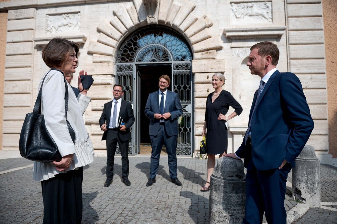 Ein Mann unterhält sich mit einer Frau vor einem Gebäudeeingang. Im Hintergrund beobachten weitere Personen das Gespräch.