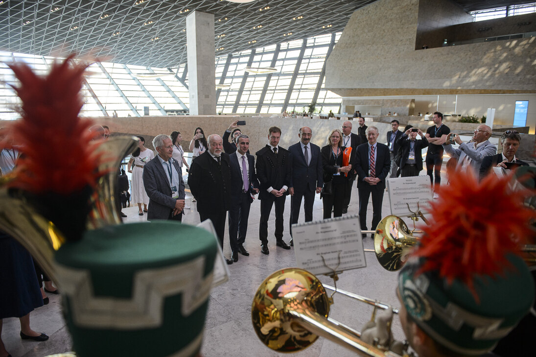 Eine Blechbläserkapelle ist im Vordergrund zu sehen. Mehrere Menschen stehen ihr gegenüber.