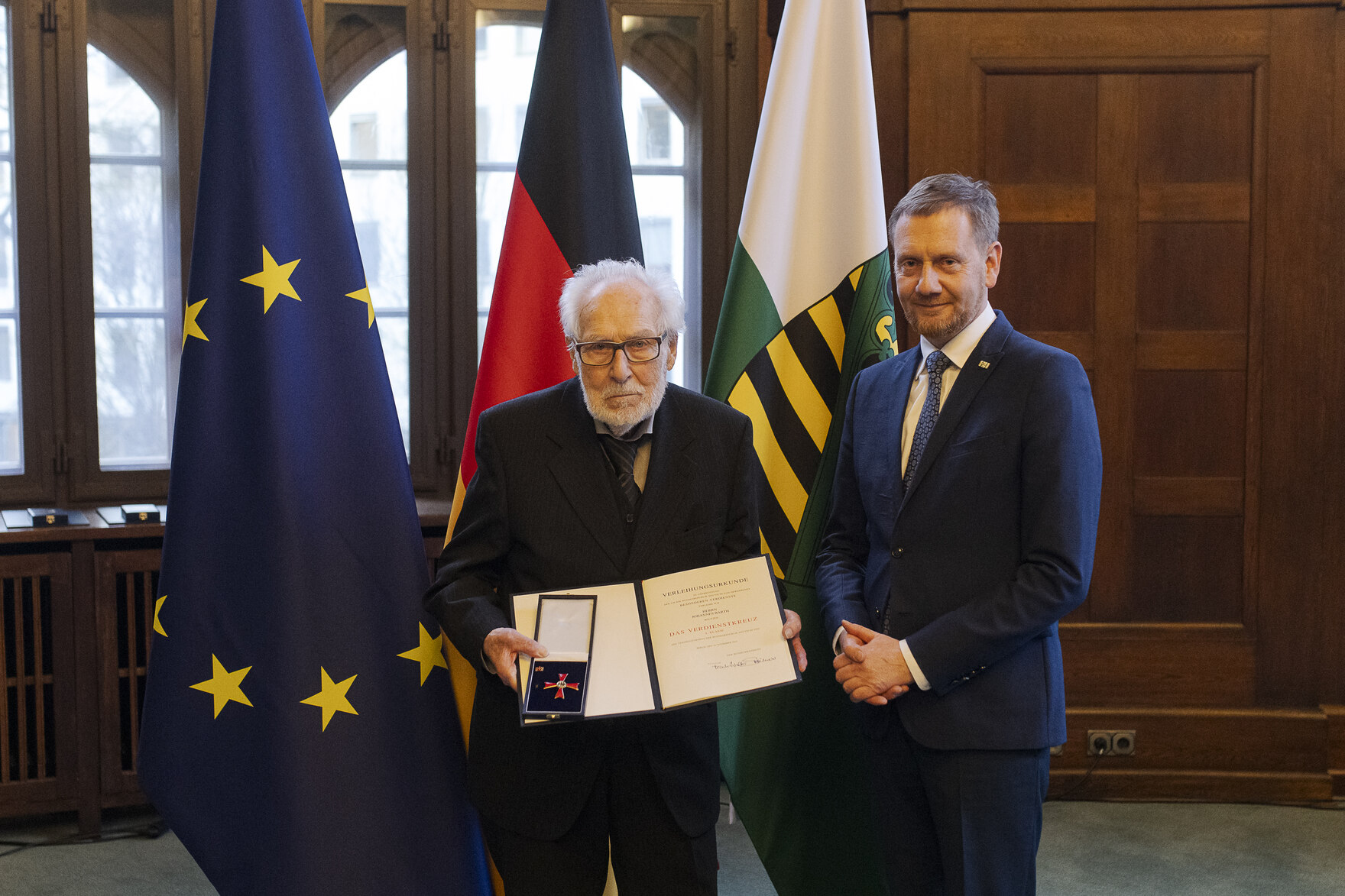 Zwei Männer in einem Raum, der Mann links hält eine offene Präsentationsmappe mit einer Auszeichnung, im Hintergrund die Flaggen der EU, Deutschlands und Sachsens.