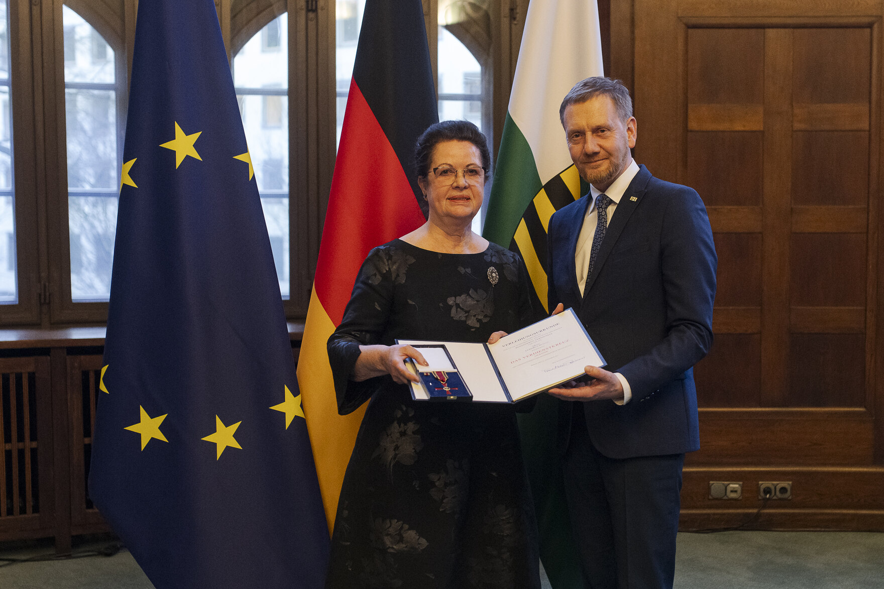 Eine Frau und ein Mann in einem Raum, die Frau links hält eine offene Präsentationsmappe mit einer Auszeichnung, im Hintergrund die Flaggen der EU, Deutschlands und Sachsens.