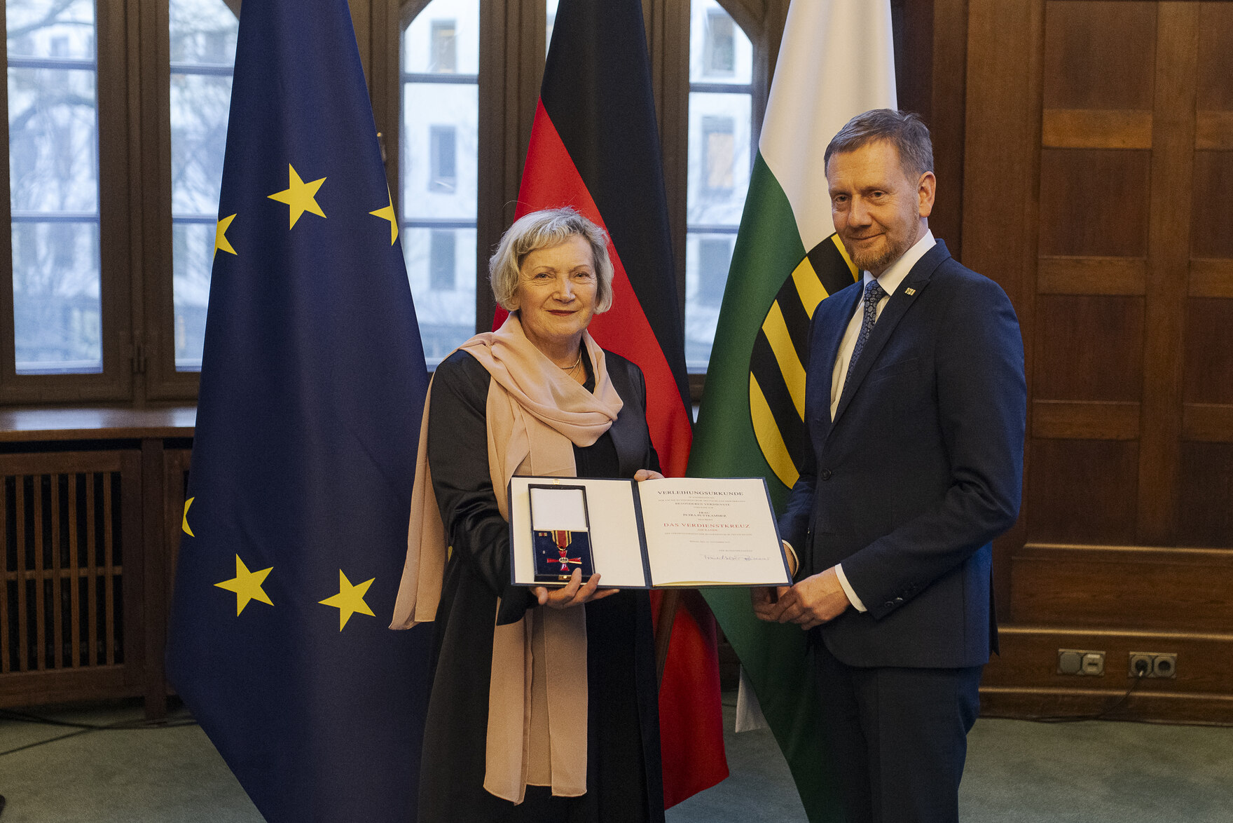 Eine Frau und ein Mann in einem Raum, die Frau links hält eine offene Präsentationsmappe mit einer Auszeichnung, im Hintergrund die Flaggen der EU, Deutschlands und Sachsens.