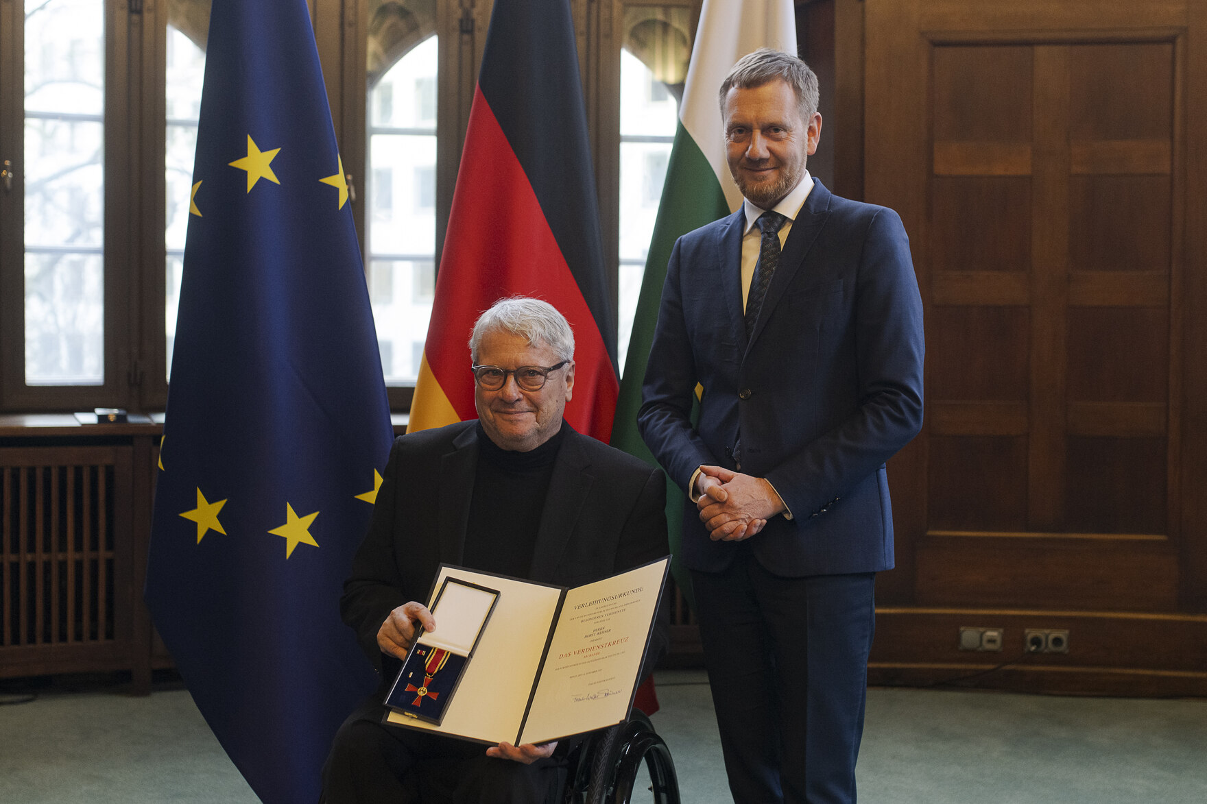 Zwei Männer in einem Raum, der Mann links (im Rollstuhl sitzend) hält eine offene Präsentationsmappe mit einer Auszeichnung, im Hintergrund die Flaggen der EU, Deutschlands und Sachsens.