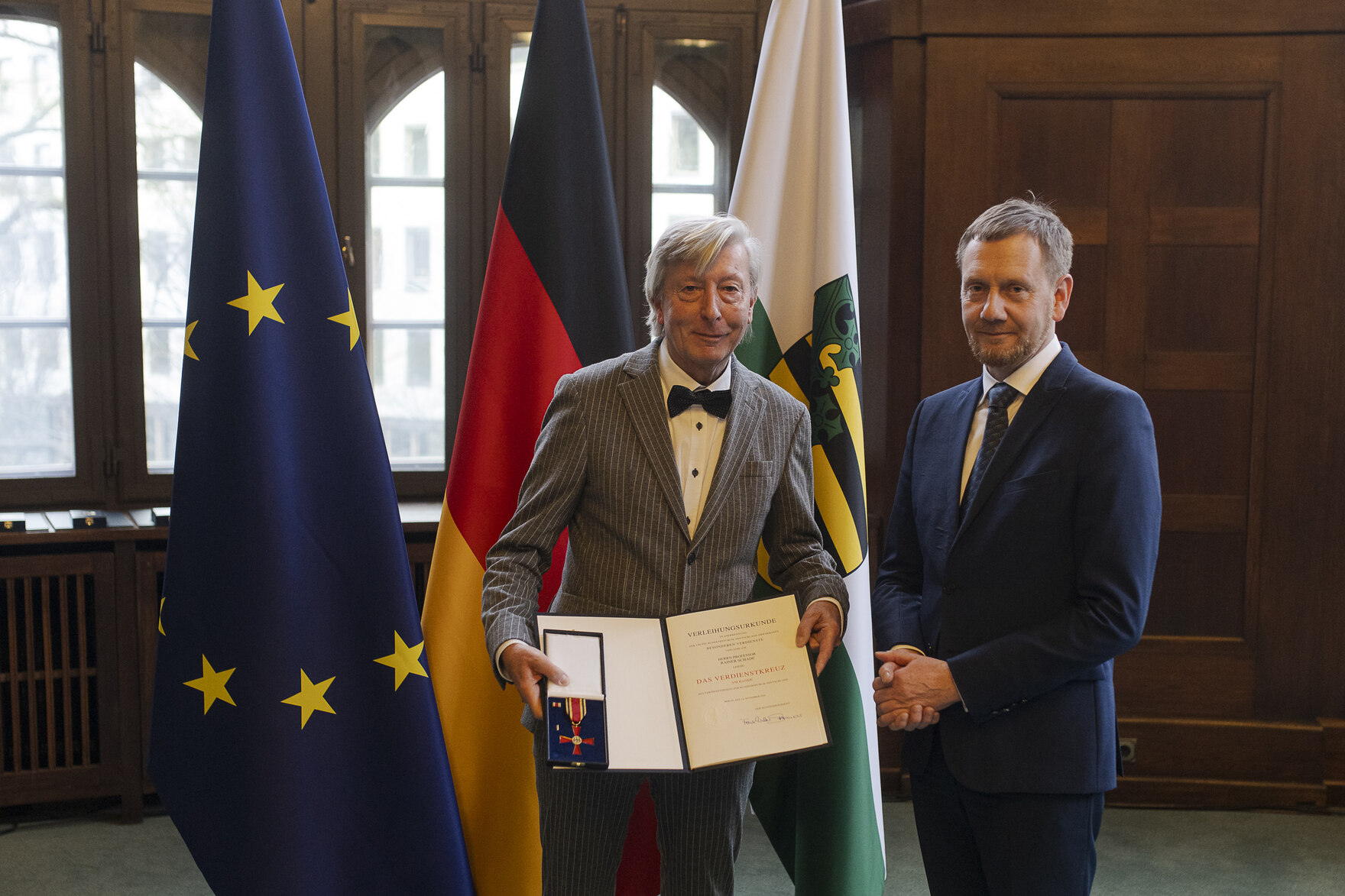 Zwei Männer in einem Raum, der Mann links hält eine offene Präsentationsmappe mit einer Auszeichnung, im Hintergrund die Flaggen der EU, Deutschlands und Sachsens.