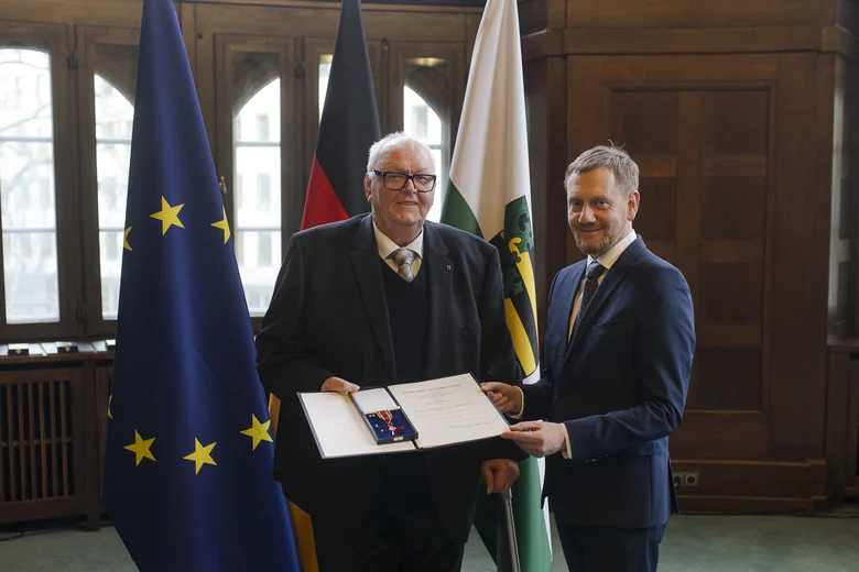 Zwei Männer in einem Raum, der Mann links hält eine offene Präsentationsmappe mit einer Auszeichnung, im Hintergrund die Flaggen der EU, Deutschlands und Sachsens.
