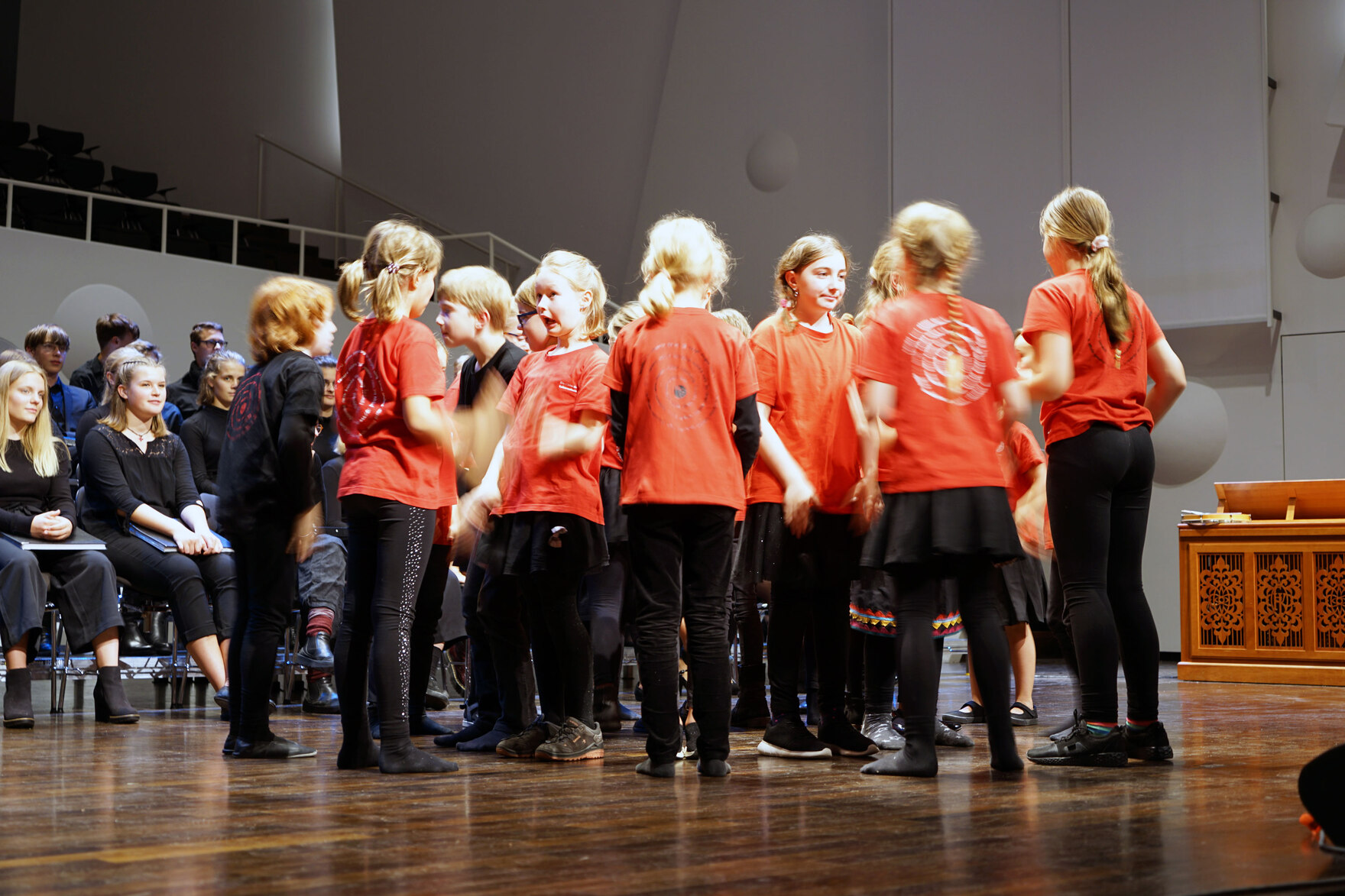 Mehrere Kinder mit roten T-Shirts stehen in einem Kreis. Im Hintergrund ist Publikum zu sehen.