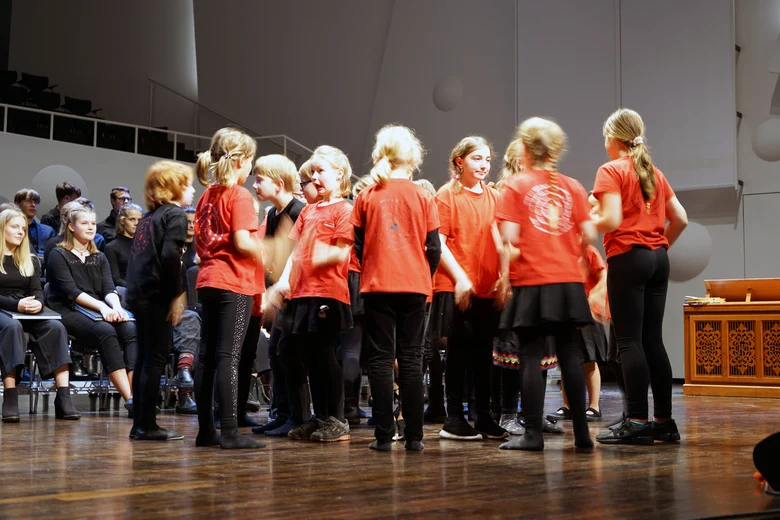 Mehrere Kinder mit roten T-Shirts stehen in einem Kreis. Im Hintergrund ist Publikum zu sehen.