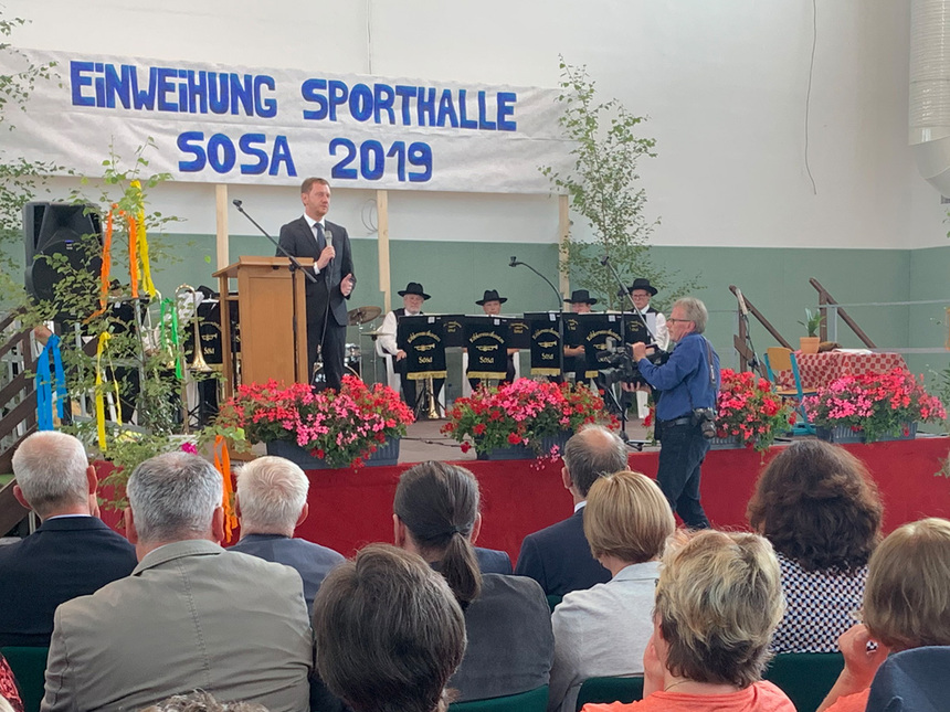 Ministerpräsident Michael Kretschmer auf der Einweihnungsfeier der neuen Sporthalle in Sosa. Auf einer Bühne steht ein Mann. Vor ihm sitzen viele Menschen im Publikum. Hinter ihm hängt eine Banner mit der Aufschrift: »Einweihung Sporthalle Sosa 2019«