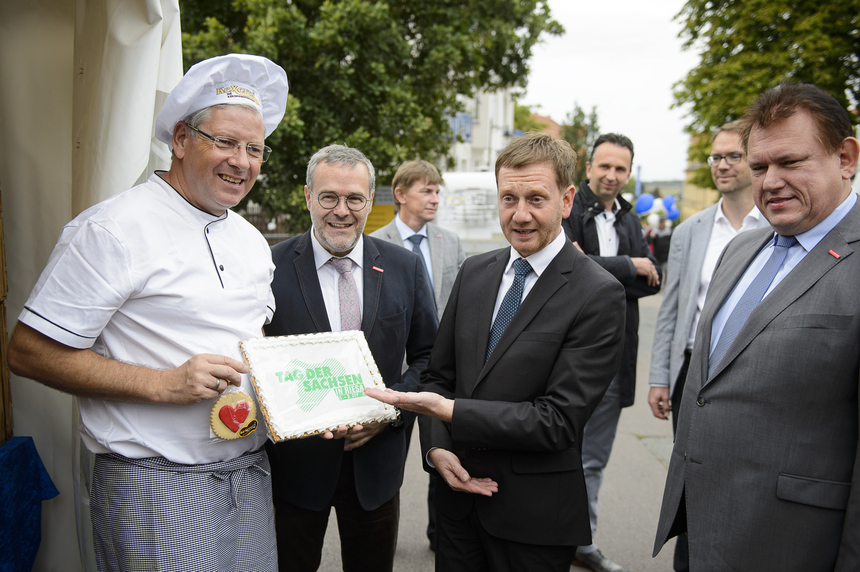 Ein Mann zeigt auf einen großen Keks mit der Aufschrift, »Tag der Sachsen in Riesa« das von einem anderen Mann in Bäckerkleidung gehalten wird.