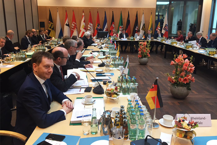 Männe rund Frauen sitzen in einem Konferenzraum an Tischen in L-Form. Im Hintergrund stehen Fahnen.