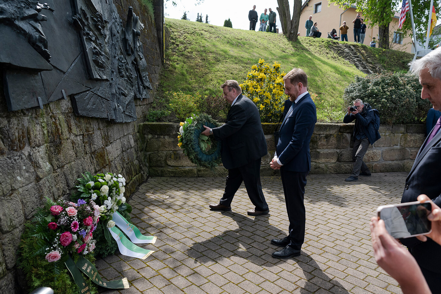 Zwei Männer stehen vor einem Denkmal. Einer legt einen Kranz nieder.
