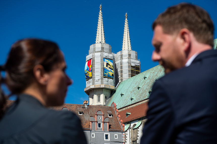 Ein Mann und eine Frau betrachten aus der Ferne einen Kirchturm, an dem Graffiti hängen.