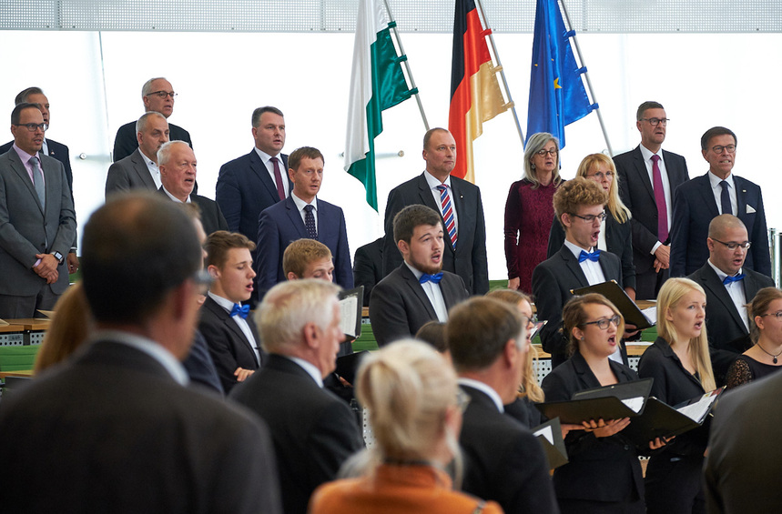 Männer und Frauen stehen in einem Saal und singen.