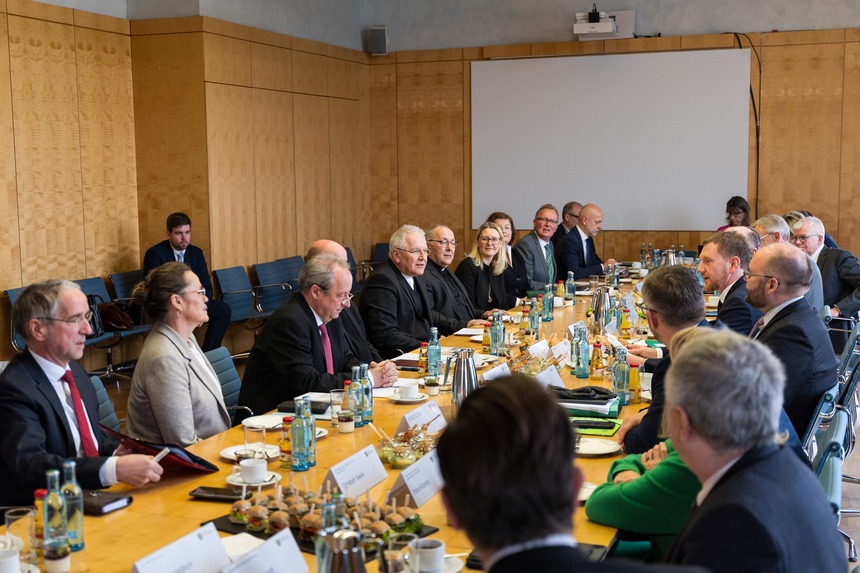 Männer und Frauen sitzen in einem Saal an einem Konferenztisch.