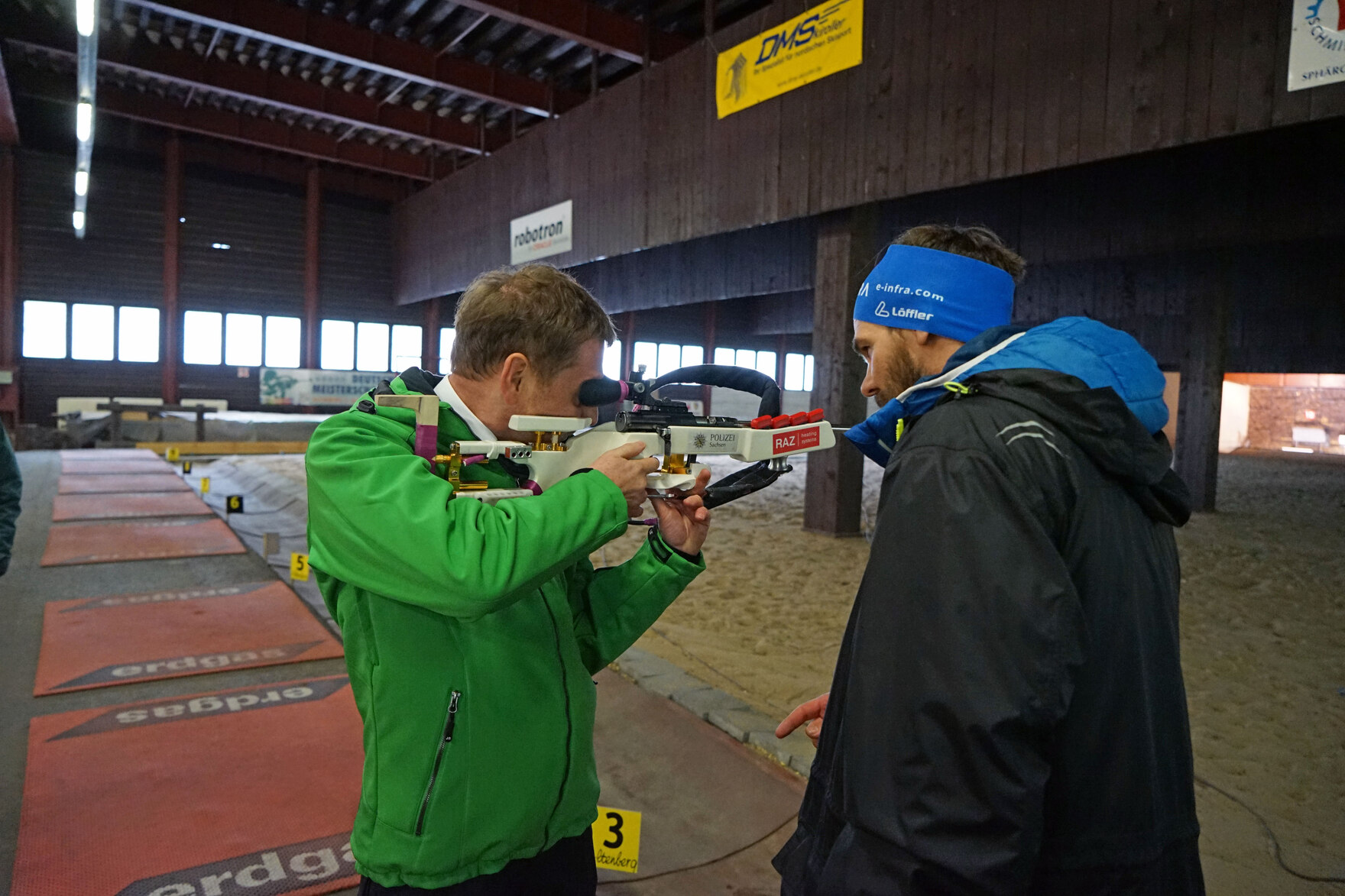 Ein Mann hält ein Biathlongewehr in den Händen während ihm ein anderer Mann anweist.