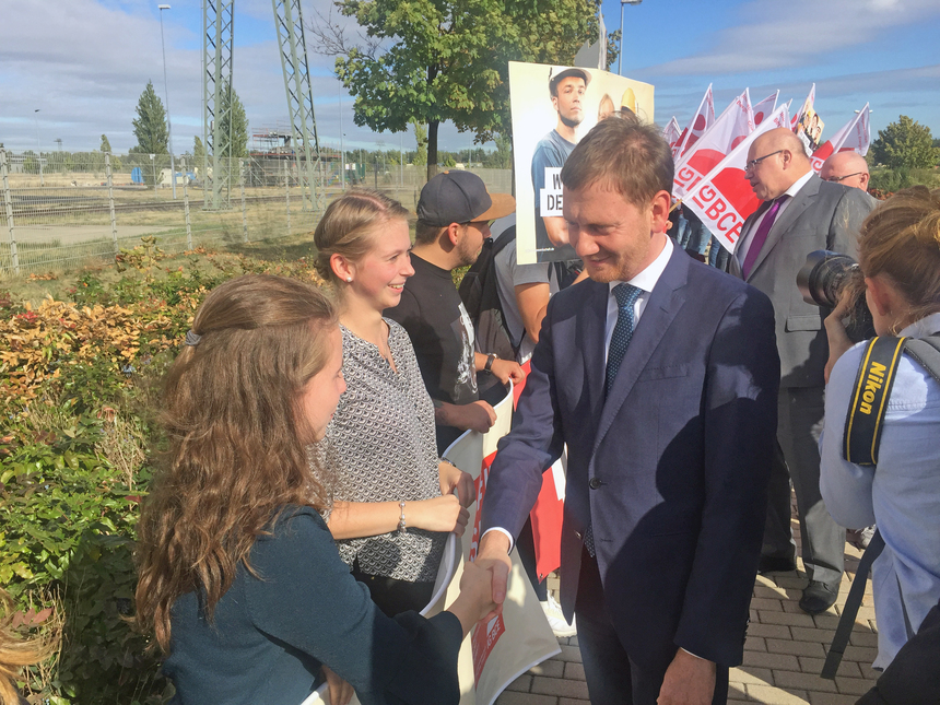 Zwei Männer sprechen mit jungen Frauen vor den Toren eines Kraftwerkgeländes.