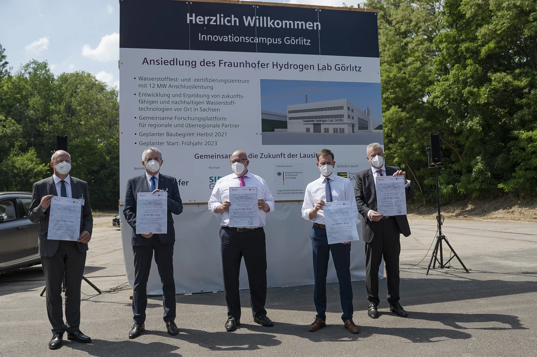 Männer stehen vor einem Bauschild und halten einen Förderbescheid in der Hand.