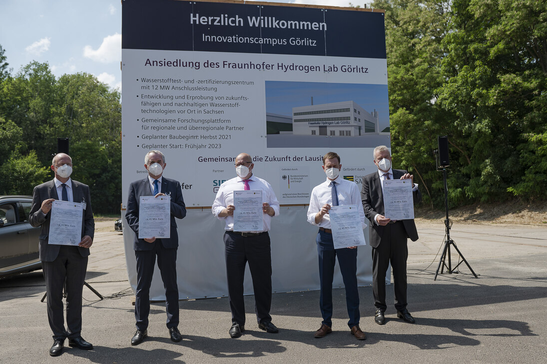 Männer stehen vor einem Bauschild und halten einen Förderbescheid in der Hand.