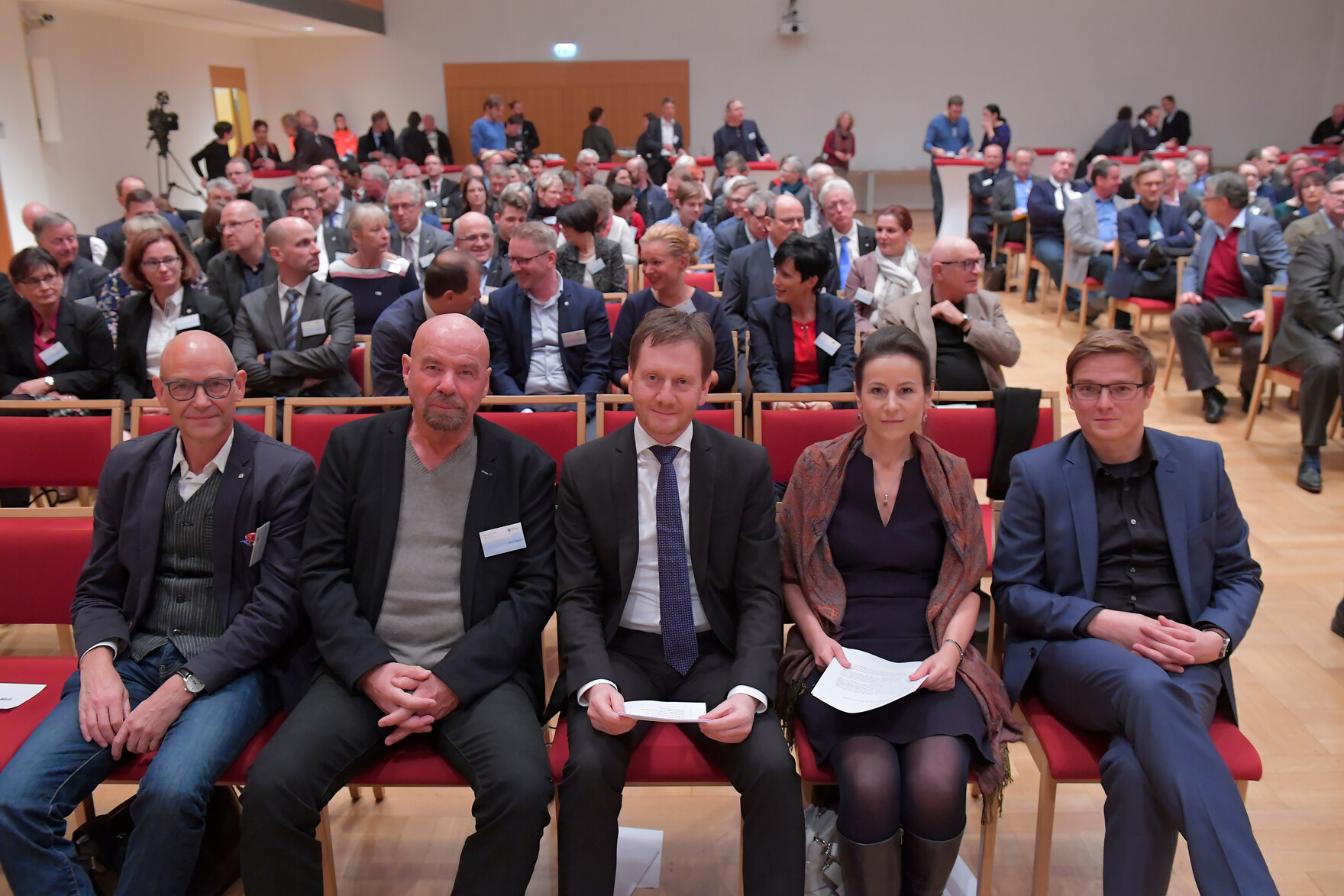 Drei Männer und eine Frau sitzen in der ersten Stuhlreihe in einem Saal.