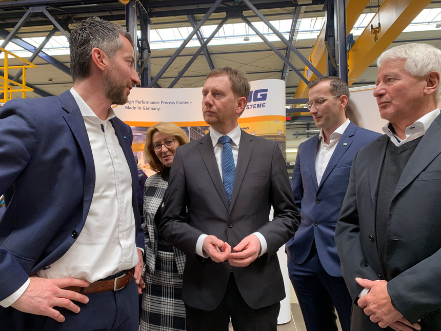 Männer unterhalten sich in einer Montagehalle. Im Hintergrund steht eine Frau.