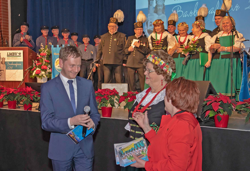 Ein Mann unterhält sich mit zwei Frauen. Im Hintergrund befindet sich eine Bühne, auf der Menschen in Bergmannskleidung stehen.