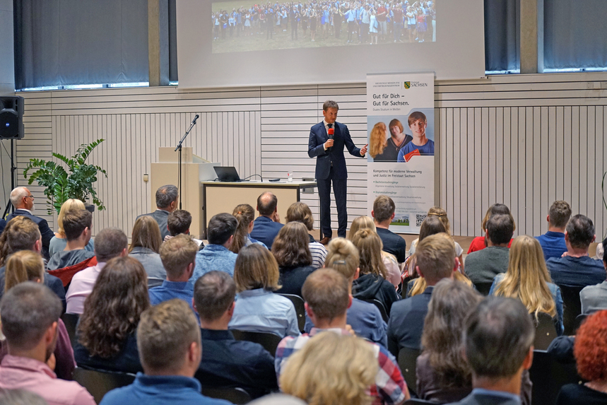 Ein Mann steht mit einem Mikrofon in der Hand auf einer Bühne. Davor sitzt ein Publikum.