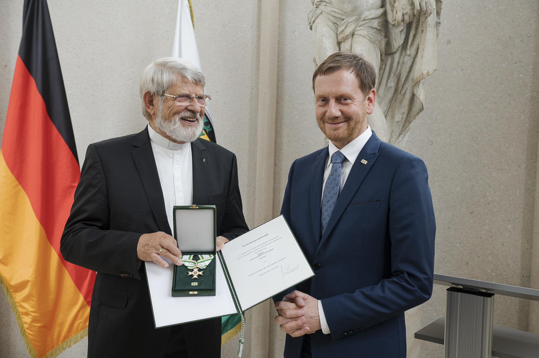 Zwei Männer stehen vor einer Fahnengalerie. Ein Mann hält eine Urkunde und einen Orden in der Hand.