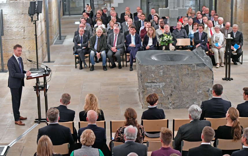 Menschen sitzen im Keller einer Kirche auf Stühlen und hören einem Mann an einem Rednerpult zu.