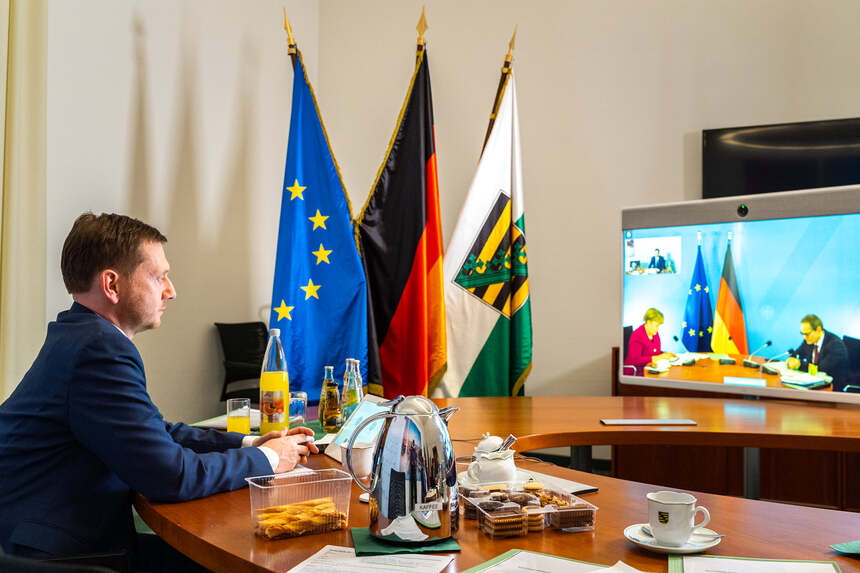 Ein Mann sitzt an einem Tisch vor einem Monitor. Im Hintergrund steht eine Fahnengalerie.