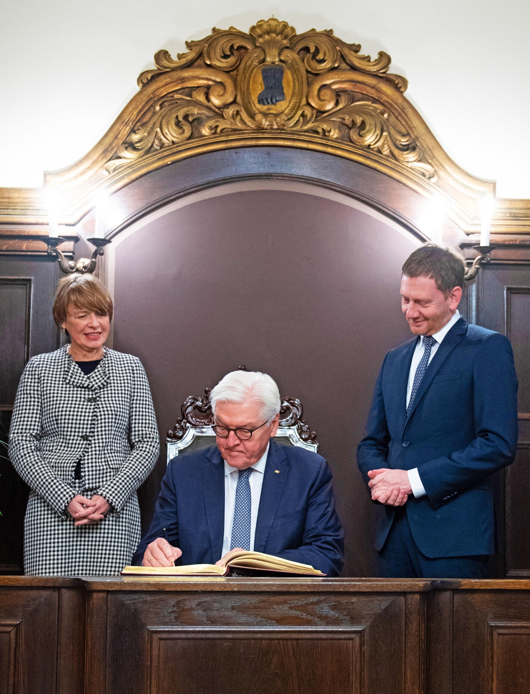 Ein Mann und eine Frau beobachten einen Mann bei der Eintragung in ein goldenes Buch.