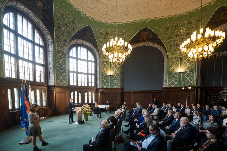 Männer und Frauen sitzen in einem Saal. Vor ihnen steht ein Mann an einem Rednerpult.