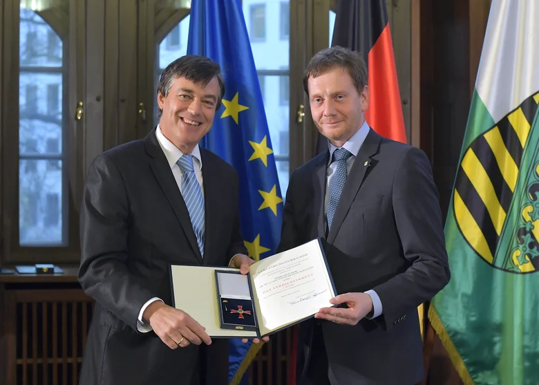 Zwei Männer stehen vor einer Fahnengalerie und halten einen Orden in der Hand.