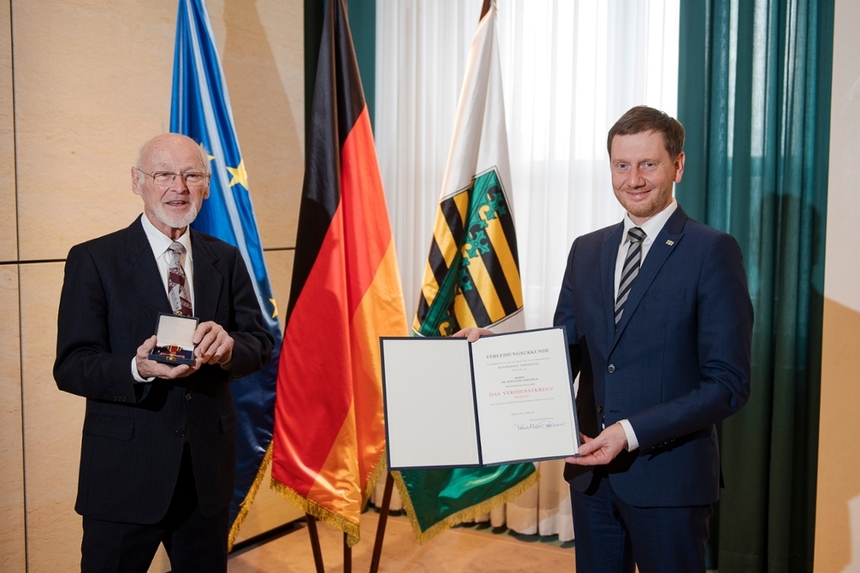 Zwei Männer stehen vor einer Fahnengalerie. Einer von ihnen hält eine Ernennungsurkunde in der Hand.