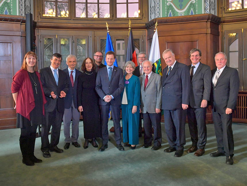 Männe rund Frauen stehen nebeneinander in einem großen Saal. Im Hintergrund steht ein Fahnenständer.