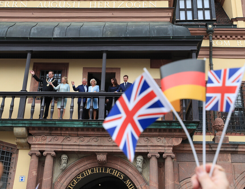 Männer und Frauen stehen auf einem Balkon. Davor wedeln britische und eine deutsche Flagge.