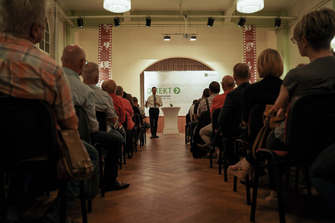 Männer und Frauen sitzen in einem Saal und hören einem Mann am Mikrofon zu.