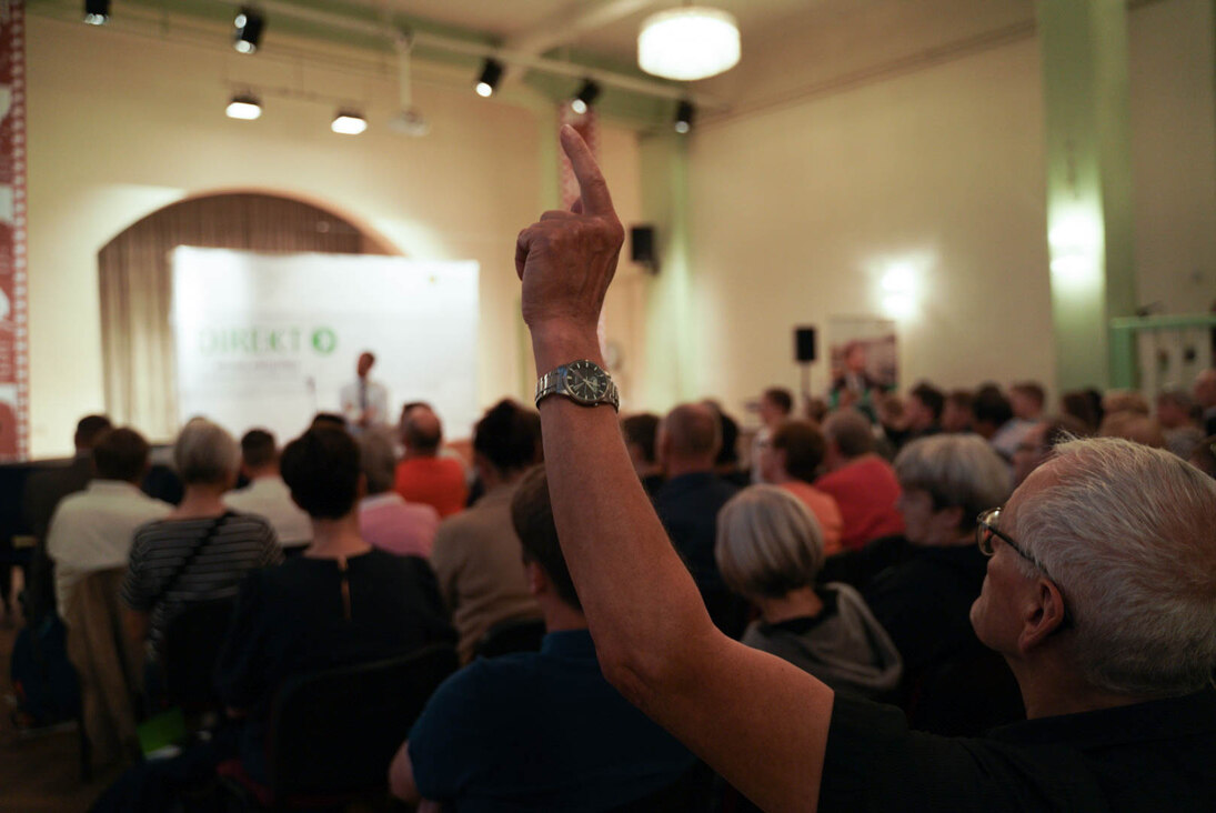 Ein Mann hebt die Hand. Vor ihm sitzen Menschen im Saal.