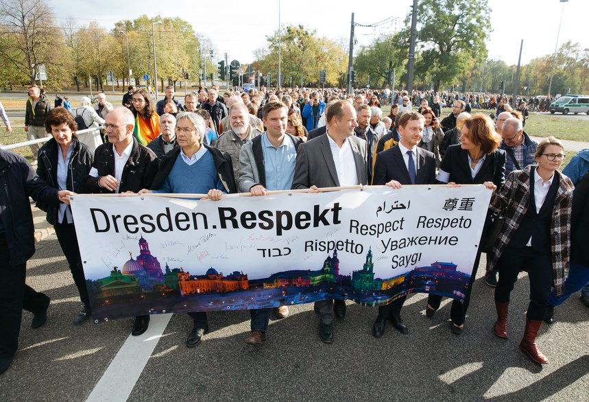 Männer und Frauen halten ein Transparent in der Hand und laufen über eine Straße. Hinter ihnen laufen viele weitere Menschen.