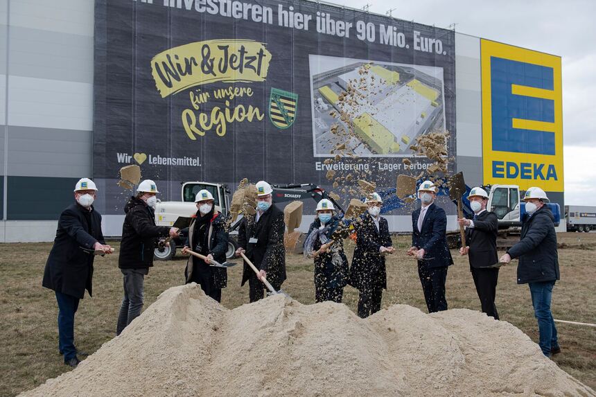 Männer und Frauen stehen vor einer großen Halle und schaufeln Sand in die Höhe.