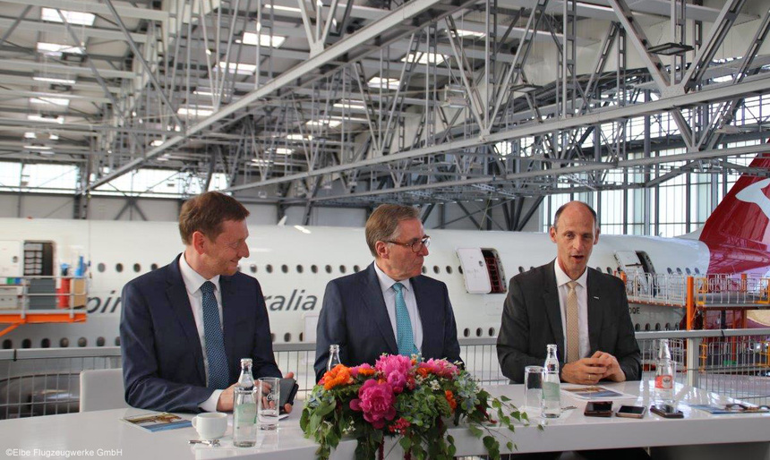 Drei Männer sitzen an einem Hochtisch. Im Hintergrund steht ein Flugzeug.