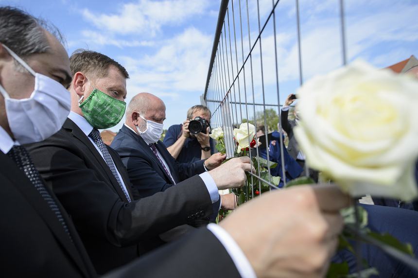 Männer befestigen Blumen an einem Bauzaun. Dabei tragen sie Masken im Gesicht