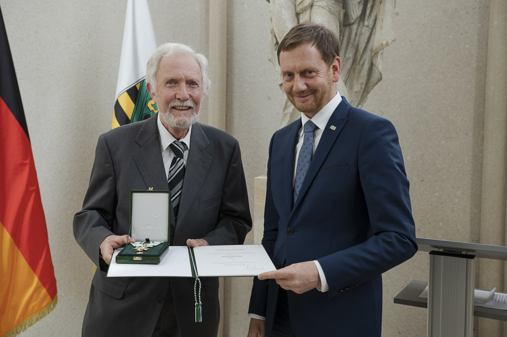 Zwei Männer stehen vor einer Fahnengalerie. Einer von ihnen hält einen Orden in der Hand.