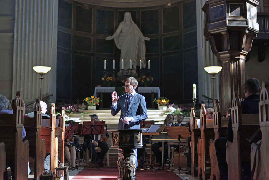 Ein Mann steht vor dem Alter einer Kirche an einem Mikrofon. Hinter ihm sitzen Musiker.