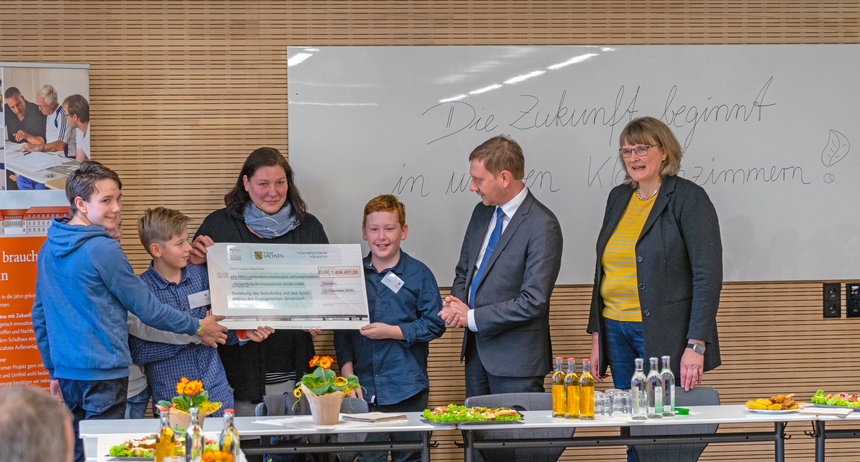 Männer und Frauen stehen in einem Klassenzimmer neben Kindern, die einen großformatigen Bankscheck in der Hand halten.