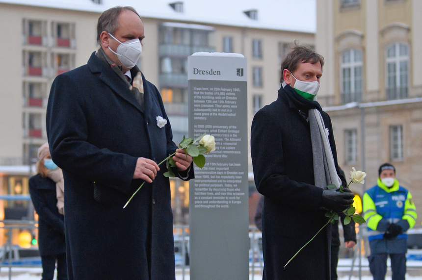 Zwier Männer stehen vor einer Stehle mit einer weißen Rose in der Hand.